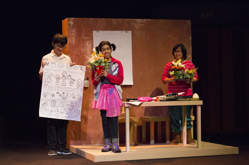 Home setting, one actor holding up a poster while the other two actors are carrying a bouquet of flowers. Photo Credit - Desmond Lui