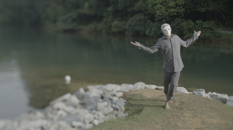 Man wearing a white mask poses with arms outstreched, standing on river shore