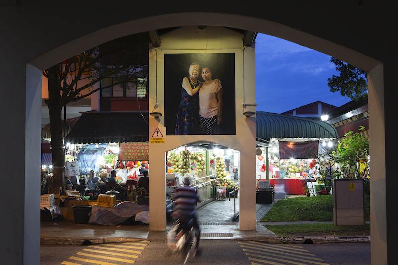 A big photo of an elderly and a young woman standing together, erected above a covered crossway near a market.