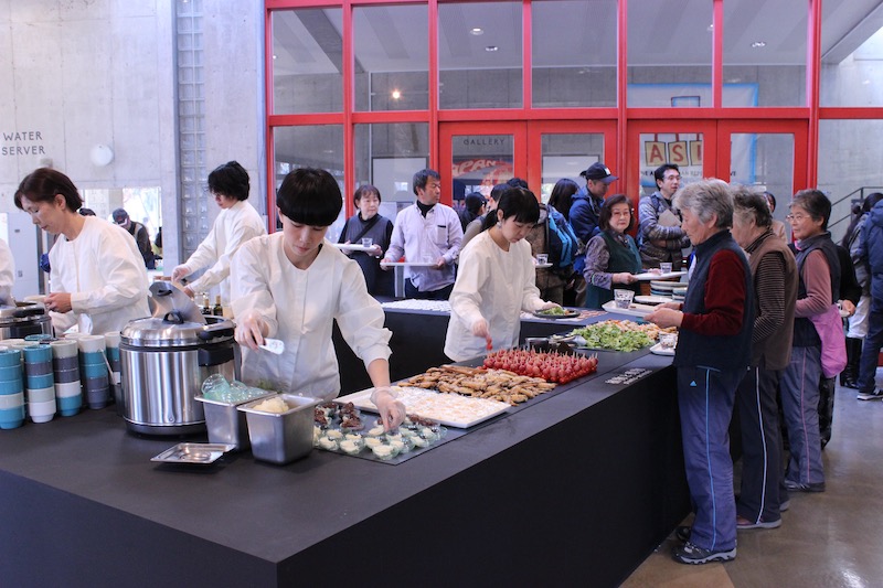 People in a canteen serving and getting served food. The servers are dressed in white.