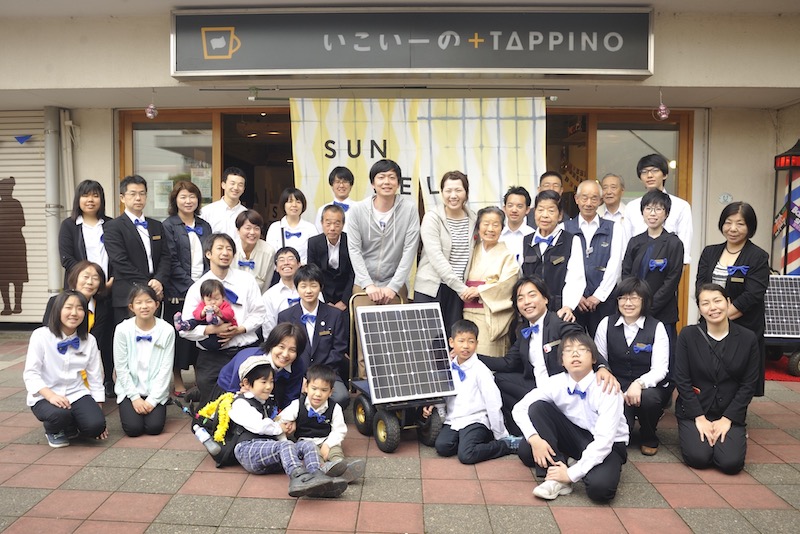 A group of people dressed in white-and-black uniform and blue bow ties posing in front of a cafe and with a solar panel in the centre.