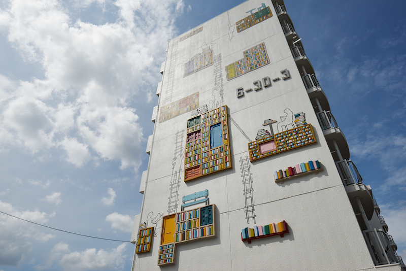 An apartment block with art fixtures of bookshelves stuck to its side. There are outlines of people reading and climbing ladders alongside these.