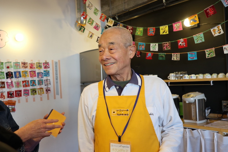 An elderly man smiling, wearing a yellow apron.