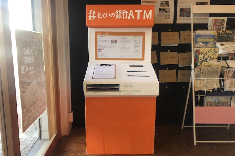 A orange-and-white cardbox box shaped like an ATM. On wall behind are booklets hanging.