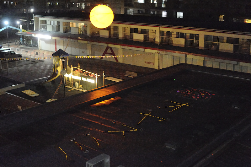 A rooftop in foreground with Japanese words made out of glowsticks on top. In background an art installation. A yellow globe resembling the sun hangs up above the buildings.