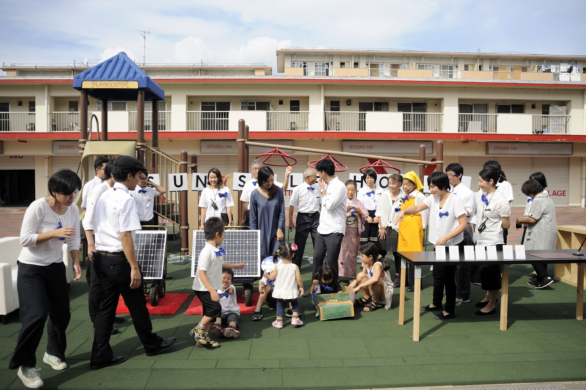 People dressed mostly in white, some with blue bowtie pins, standing in front of a lowrise building, with two solar panels.
