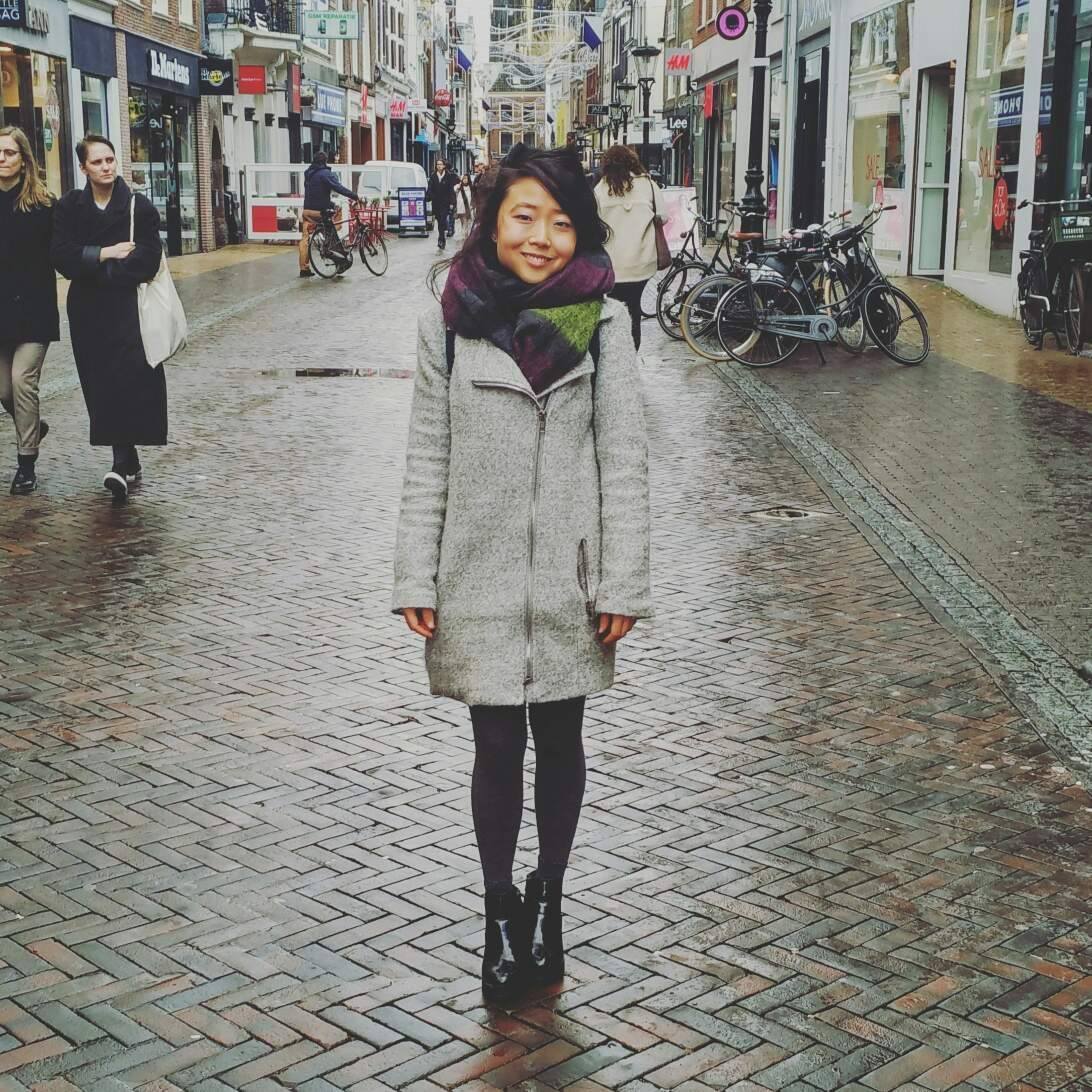 Kirin Heng is standing on a brick street that is wet with rain and wearing a winter coat and smiling.