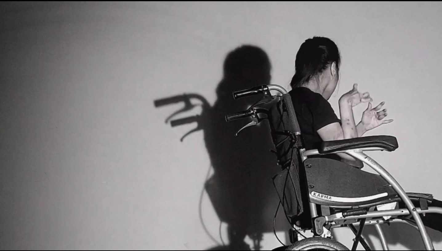 Stephanie is sitting in her wheel chair, near to a wall. The lighting is dark, casting a sharp shadow of her against the wall next to her.