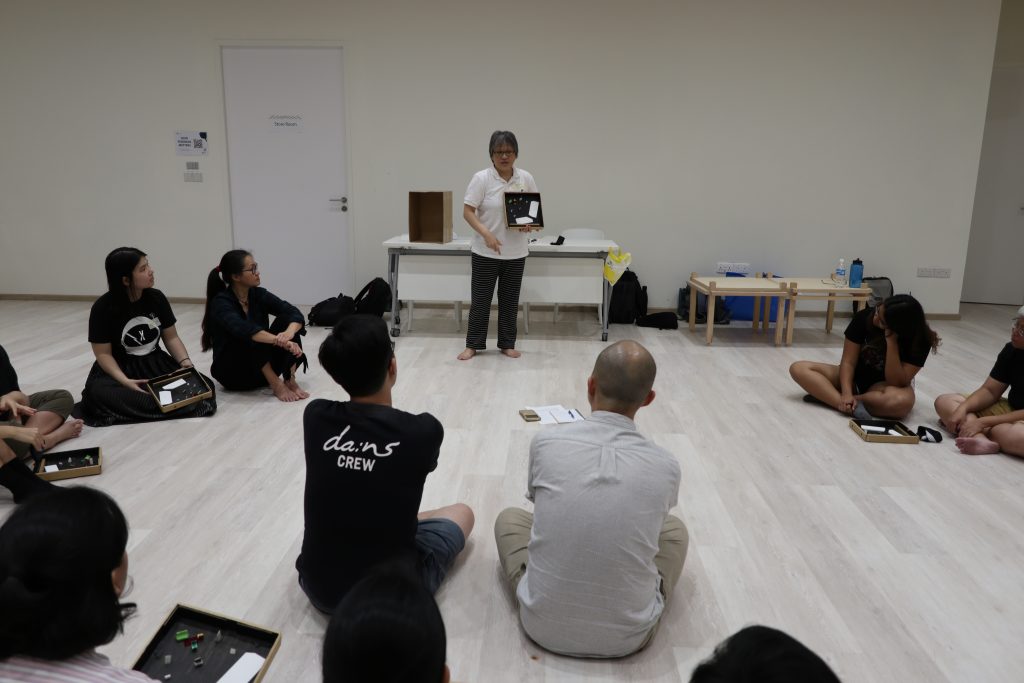 Tan Beng Tian speaking to a circle of volunteers seated on the ground. She is standing and holding a tactile board.