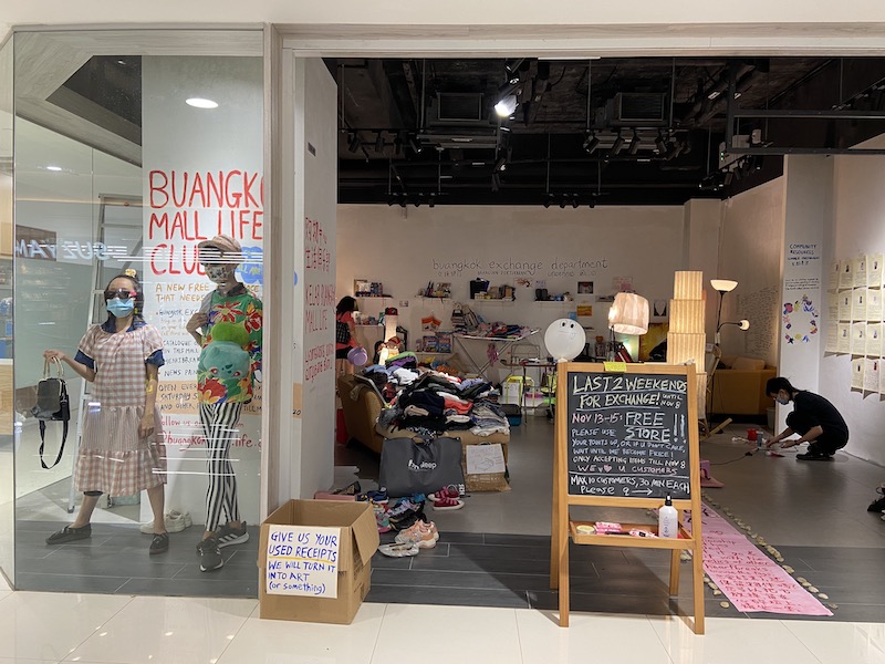 (From left) Shop assistants Crystal Ng and alex t. are modeling in the glass window of the store, dressed in clothes and accessories donated to the store. Behind them is the mural wall, with the words “Buangkok Mall Life Club” painted in capital letters with red paint. Inside the store, shoes, clothes, furniture, and various items can be seen arranged and clustered around the space. At the back of the store, the words “ buangkok exchange department” are written on the wall, below it is the Chinese, Malay, and Tamil translations. The right wall is plastered with “posters” of customers who have visited the store. At the entrance (from left), a piece of paper that reads “GIVE US YOUR USED RECEIPTS WE WILL TURN IT INTO ART (or something)” is stuck on a small cardboard box. Next to it is a small chalkboard that reads “ LAST 2 WEEKENDS FOR EXCHANGE! Until Nov 8, Nov 13-15: FREE STORE!!