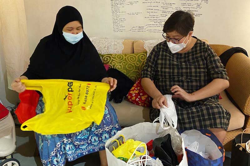 (From left) Linda is in a black tudong and blue floral dress, holding a yellow plastic bag. Diana is in a brown checkered dress, wearing black glasses and folding a white plastic bag Both customers are seated on a light brown sofa, folding plastic bags into a big white paper bag in front of them.