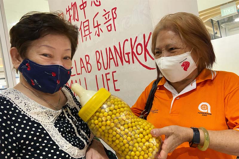 Two senior ladies holding a giant milk bottle containing sweets. Behind them is the sign for the Buangkok Mall Life Club.