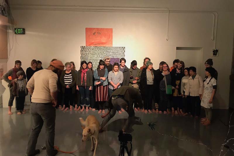 Background: participants lining up against the wall, barefooted Foreground: (left) guy in beanie facing the participants while looking at the dog, (center) a dog on a leash Panelists, (right) guy in black jacket bending down to take the photo probably