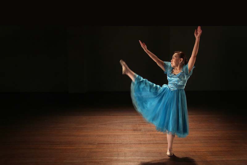 Koh Lian Hiok in light blue ballet tutu dress, hair tied up in a bun. The photo captures her in mid-dancing, where she is posing with her right leg pointed upwards and both her hands elegantly raised.