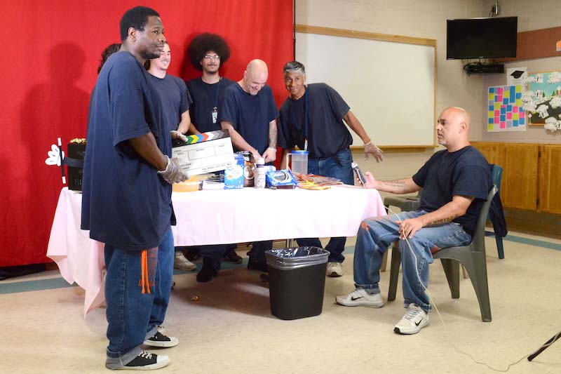 Inmates are all in navy blue t-shirts and jeans, crowding in front of a red backdrop and around the table of the Microwave Magic set, making a chocolate cheesecake. David holding onto the clapperboard while Irvin Hines seats on a chair holding onto the microphone/recorder