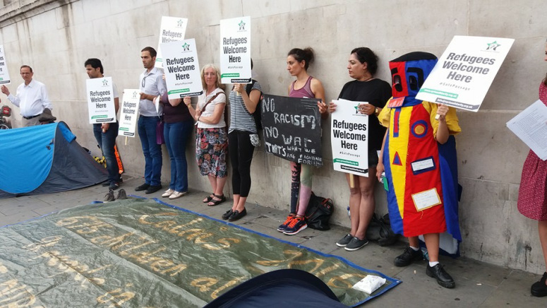 A line of people holding signs and standing against a wall. The signs mostly say "Refugees welcome here." Fié is wearing a colourful red and yellow costume.