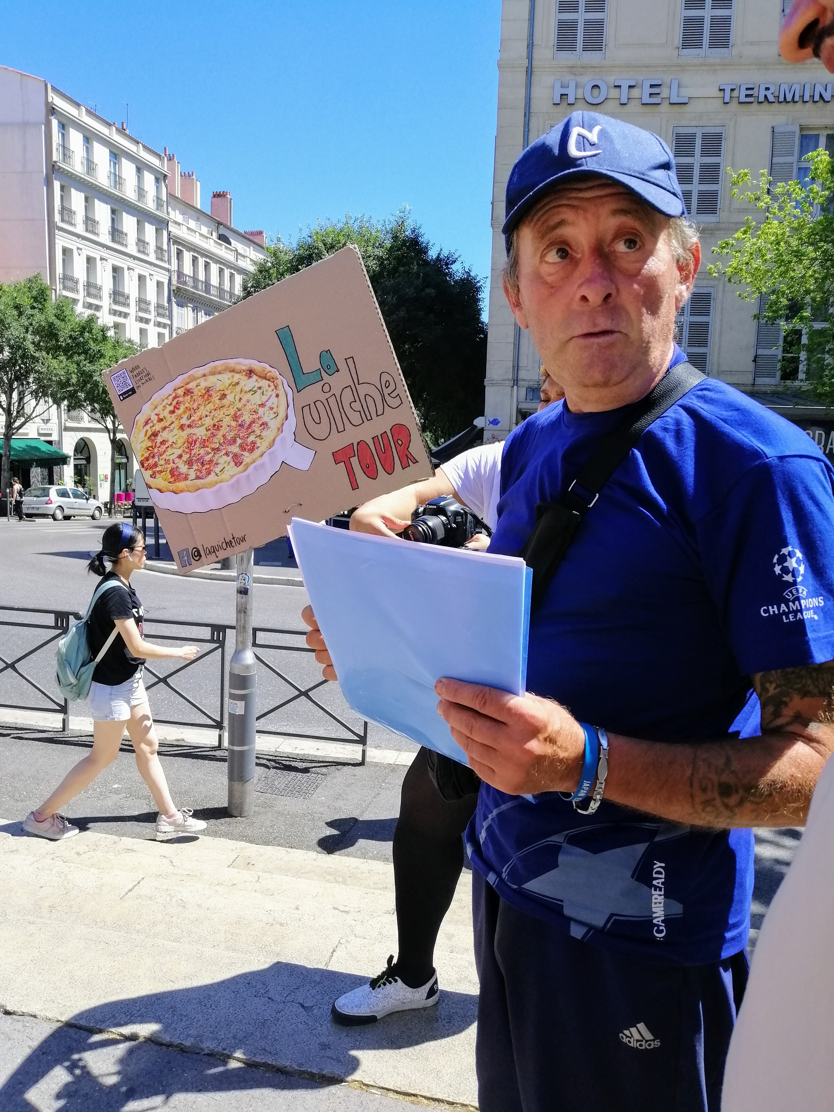 A man with a cap on, standing outdoors with a sign with words "La Quiche Tour", which has a picture of a quiche on it too.