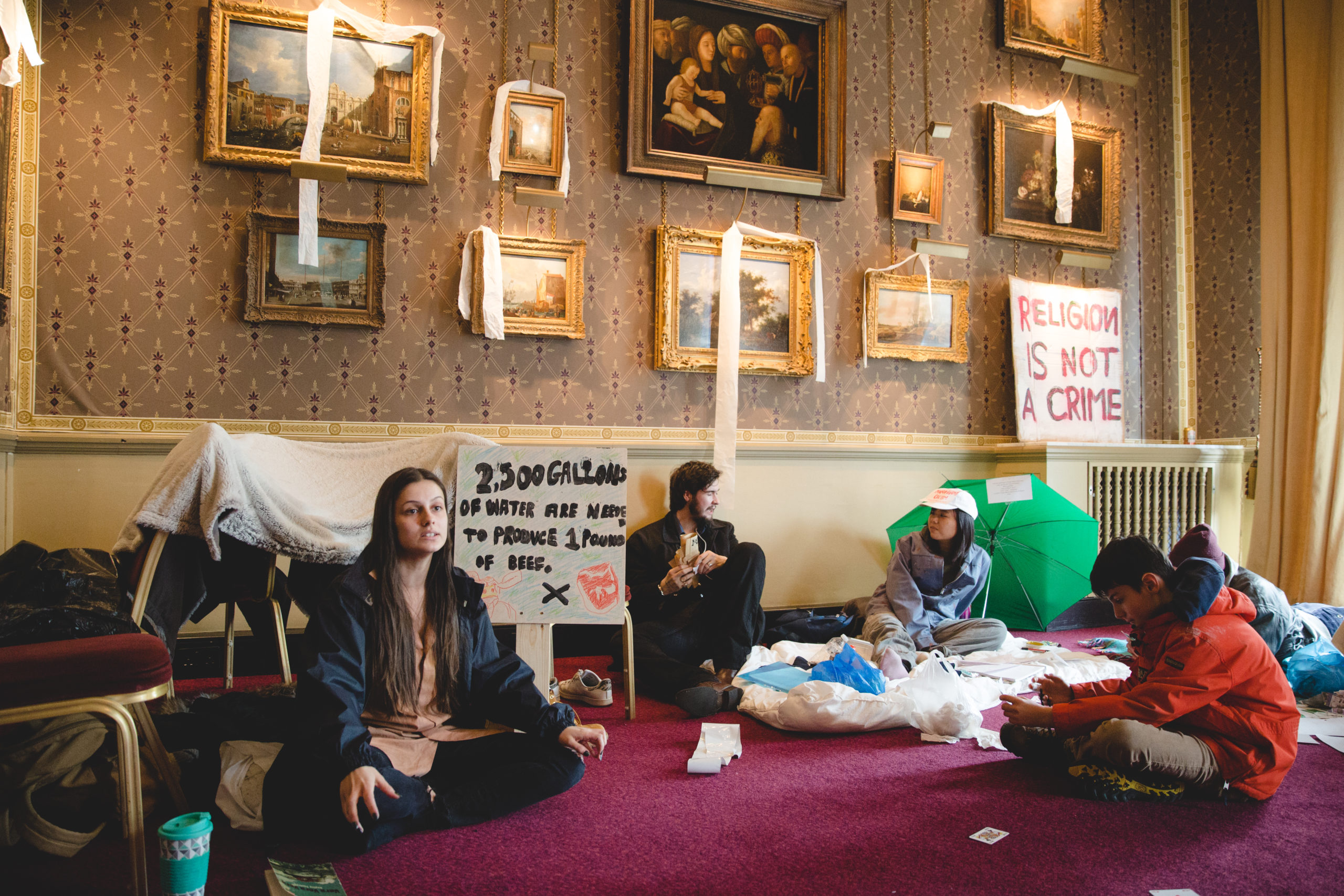 Students sitting in a museum gallery room of fine art paintings, with signs that bear messages such as "religion is not a crime"
