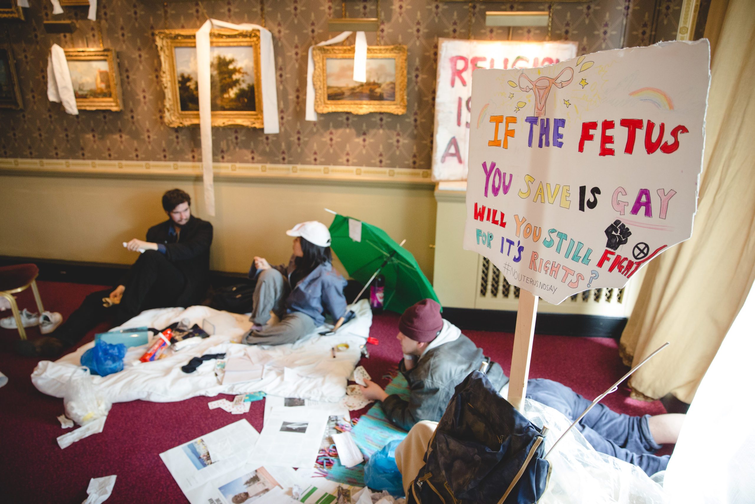 Students sit on the floor of a museum room with paintings on the walls. There is a large sign behind them with the words "If the foetus you save is gay, will you still fight for its rights?"