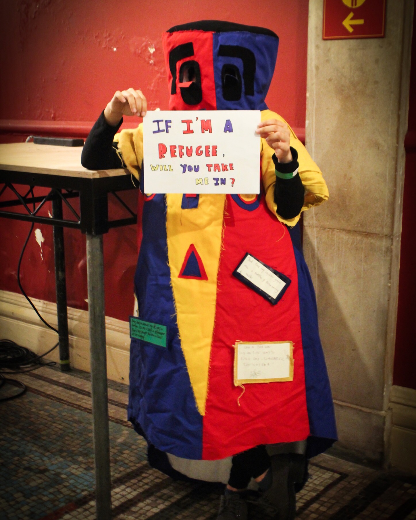 Fie wearing a red and yellow costume that obscures her face, holding up a sign which says "if I'm a refugee, will you take me in?"