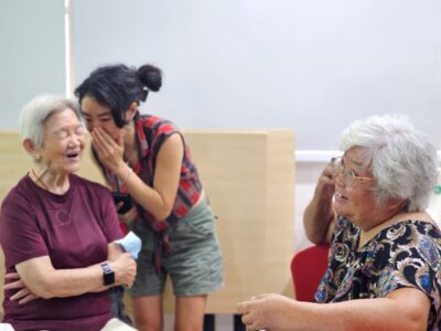 Artswok Both Sides, Now Yishun Seniors Conversations with artist Salty Xie whispering into the ear of a senior lady laughing with her eyes closed