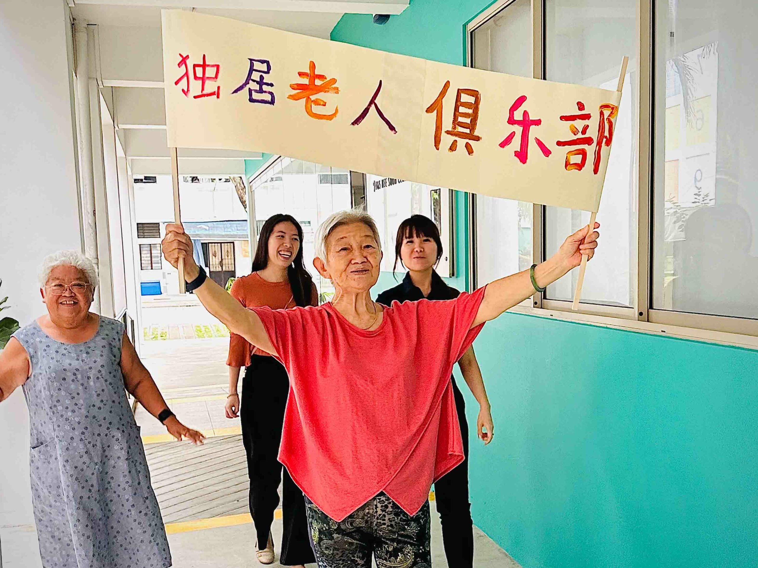 Tong Cheng holding up a banner with text in Chinese: Single Seniors’ Club as Yim Fong and 2 people follow behind. 